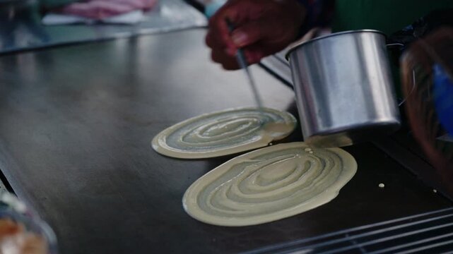 Asian street food vendor preparing thin pancake rolled crepe with sausage on hot pan.