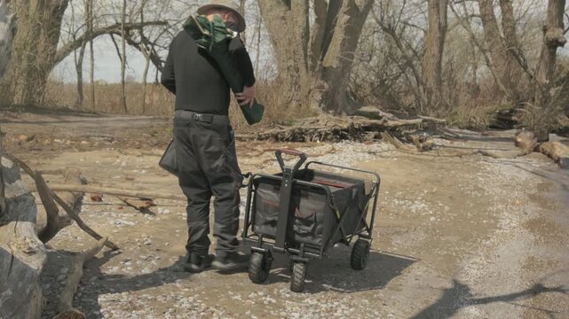Senior man packs bags and gear into utility cart on wooded lakeshore at Lynde Shores Conservation Area, Whitby, Ontario, Canada.