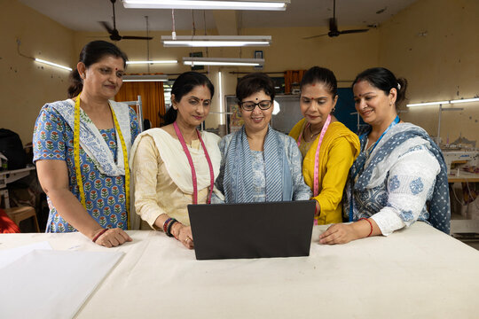 Group of skilled Indian women workers collaborating on a laptop inside a modern textile company. Female employees using digital technology for production planning, quality control and training.