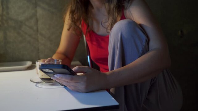 A cozy woman scrolls on her smartphone by a cup of coffee at a table in a dimly lit room. She feels calm and peaceful during this quiet moment.