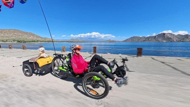 An elderly woman enjoys biking on a recumbent ebike tricycle by the lake, pulling her dog in a small trailer behind her.