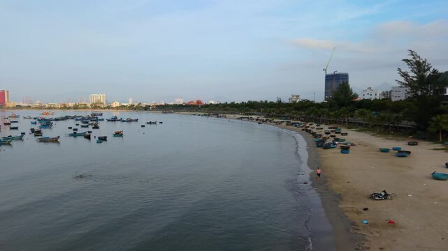 Smooth low drone flight following a quiet sandy beach with anchored fishing boats and the Da Nang skyline at sunrise.