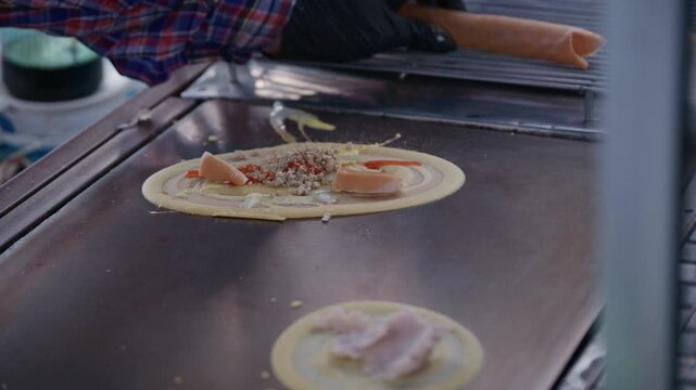 Asian street food vendor preparing thin pancake rolled crepe with sausage on hot pan.