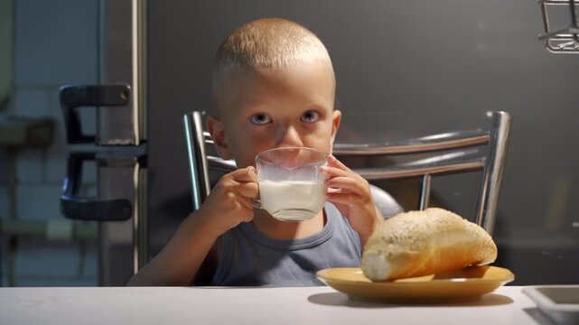 Toddler-aged boy seated at kitchen table holding glass mug of milk with both hands and sipping toward camera, with single bread roll on small plate beside him and metal chair back visible