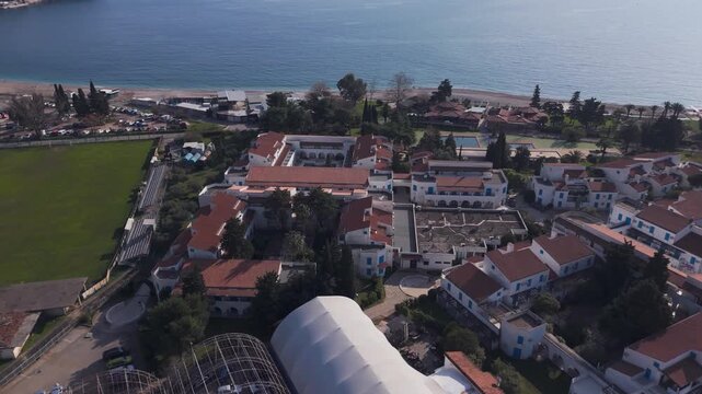 Aerial drone flyover of a luxury hotel complex with terracotta roofs and swimming pool near a green stadium, revealing the waterfront promenade and turquoise sea in Rafailovici, Montenegro.