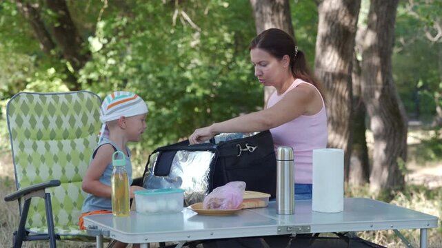 Young mother prepares baby bottle at picnic table while toddler sits in portable high chair nearby, with leafy park shade, folding chairs, picnic supplies and calm family outing atmosphere
