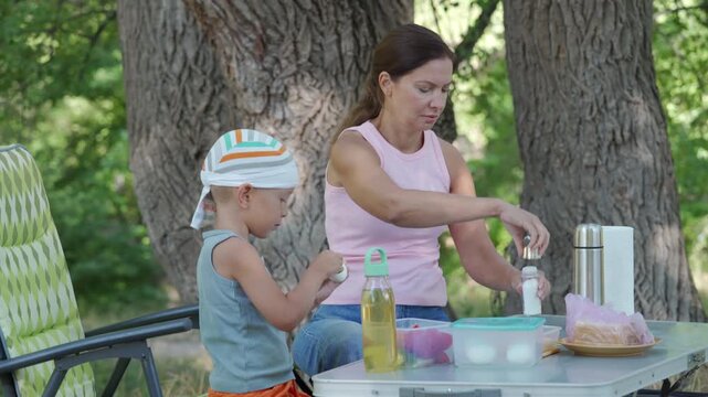 Woman and young girl arranging picnic items on metal bench beneath mature trees in shaded park setting, with striped sun hat, summer dress, shared food, cups, and relaxed family moment