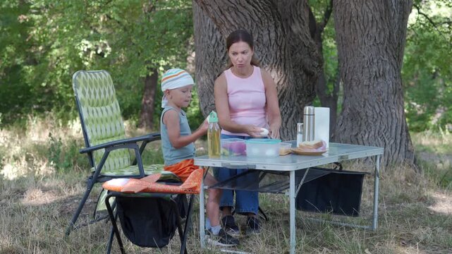 Middle-aged woman and young girl stand at portable table beneath tall shade trees, preparing drinks and snacks beside cooler and folding chairs in calm summer park scene, with woman, child, girl