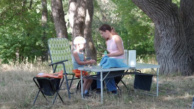 Young mother feeding toddler at picnic table beneath large oak tree in shaded park clearing, with folding chairs, cooler, snacks, dappled sunlight, green grass, relaxed summer family time
