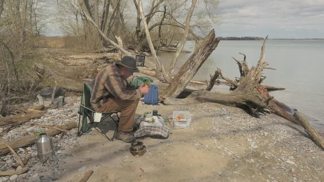 Senior man sits in camping chair eating a sandwich beside driftwood on Lake Ontario shoreline at Lynde Shores Conservation Area, Whitby, Ontario, Canada.