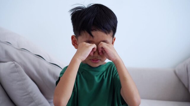 Asian boy rubbing his itchy eyes at home on a couch. Concept of pediatric health, allergy symptoms, eye fatigue, vision problems, and sleepy.