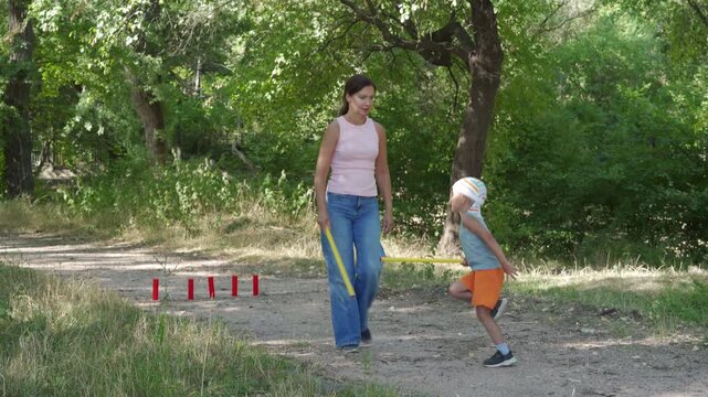A loving mother and her happy son joyfully play a game of gorodki on a dirt path. They share a cheerful, bonding moment. A peaceful green forest surrounds them.
