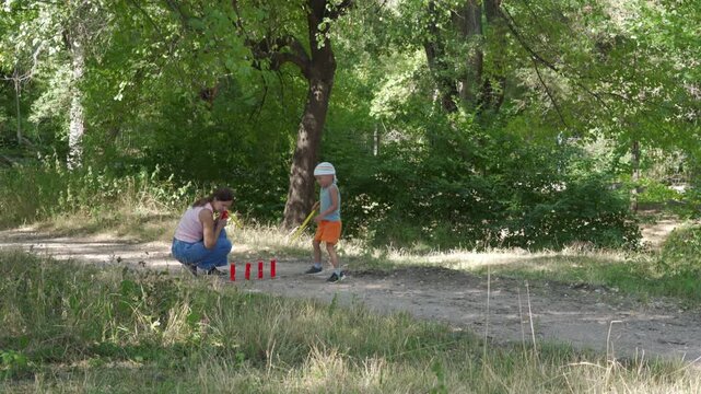 A loving mother and her happy son joyfully play a game of gorodki on a dirt path. They share a cheerful, bonding moment. A peaceful green forest surrounds them.