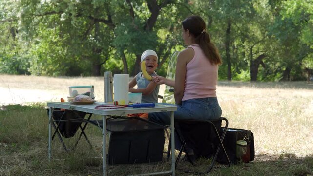 A loving mother hands a banana to her laughing son at a picnic table. They share a joyful, playful moment. A sunny, green park surrounds them.