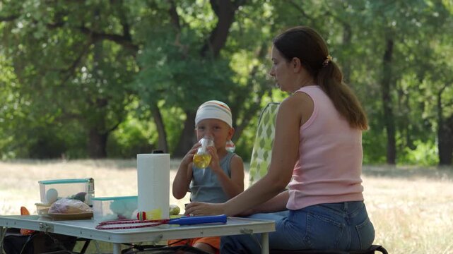 Young mother sits beside toddler at portable picnic table in shaded park clearing, child in sun hat eats snack while mother watches closely and arranges food, with dappled sunlight and leafy greenery