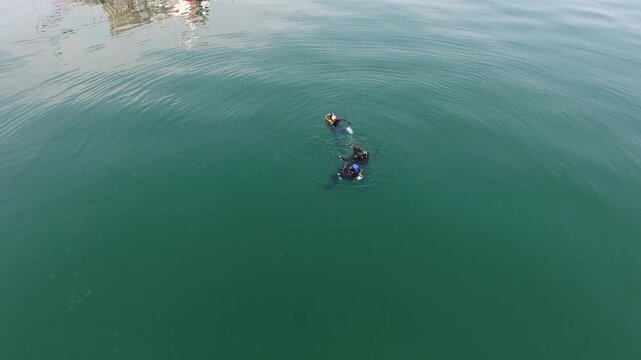 High angle aerial view of three scuba divers in wetsuits floating at the surface of calm green water beside a dive boat, with a marker buoy and rising bubbles