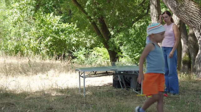 Young boy in shorts and cap walks toward portable folding table while woman stands nearby beneath large oak trees in sunlit park clearing, showing casual outdoor gathering, grassy ground, shade, and