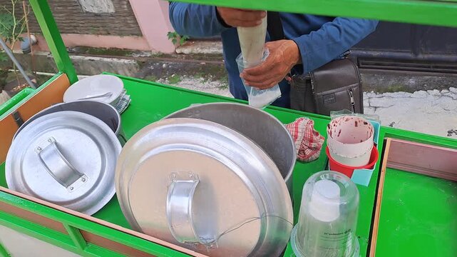 Buy mung bean porridge from a traveling vendor using a cart. There are two steamers or pots, one containing the mung bean porridge and broth, and the other containing black sticky rice. 