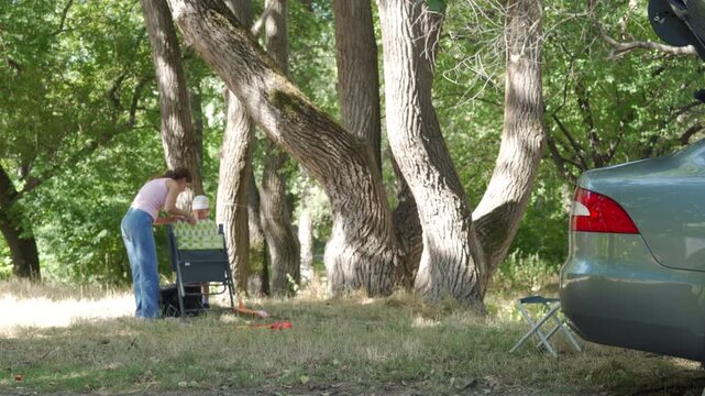 A content mother and her helpful son carefully pack up their picnic gear. They share a peaceful, satisfied feeling at the end of the day. Large forest trees and a car surround them.