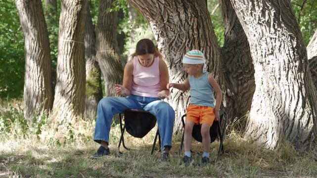 A tired mother and her curious son rest on folding chairs. They share a quiet, peaceful moment after their picnic. Large, shady forest trees surround them.