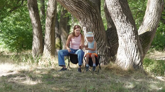 A loving mother and her curious son sit on camp stools in a peaceful forest. They smile joyfully while examining a tiny twig together.