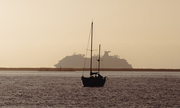 A serene image depicting a small sailboat in the foreground with a majestic cruise ship silhouetted against the hazy sunset sky, reflecting peace and adventure.