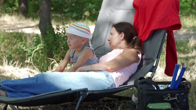 Young woman reclining on padded lounge chair while toddler sits beside her wearing headband and shorts, creating relaxed family moment in shaded grassy park with dappled sunlight, soft comfort and