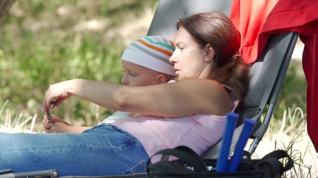 A relaxed mother and her calm son cuddle on a lounge chair. They share a loving, cozy moment while resting. A peaceful green park surrounds them.