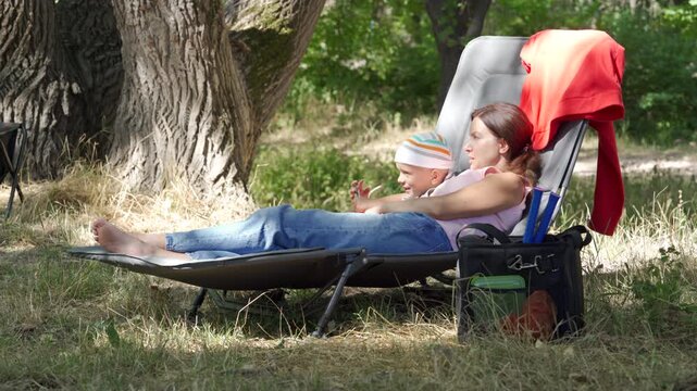 A relaxed mother and her calm son cuddle on a lounge chair. They share a loving, cozy moment while resting. A peaceful green park surrounds them.