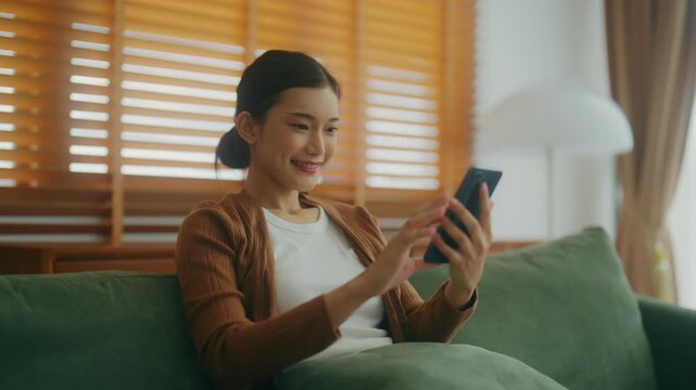Excited happy young Asian woman holding smart phone device sitting on sofa at home - Happy satisfied female looking at mobile smartphone screen and gesturing greeting friend with video call