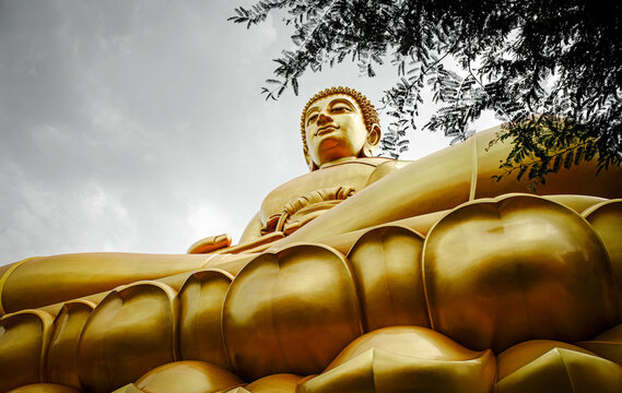 The Big Buddha at Wat Pak Nam Phasi Charoen: A low-angle view of the magnificent golden Buddha statue against a cloud-covered sky. Representing Buddhism, spirituality, peace, faith, and Thai culture.