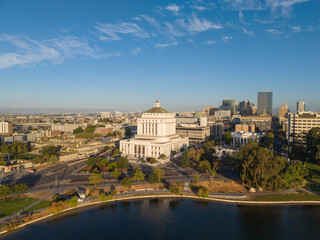 Fototapeta premium Aerial view displaying the iconic Oakland City Hall building next to Lake Merritt with the downtown skyline under a clear blue sky, highlighting urban landscape and government architecture