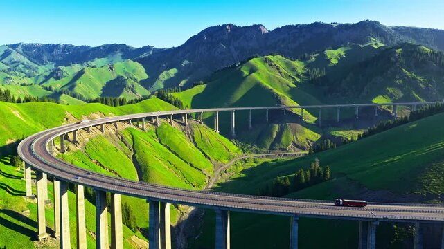 Aerial view of a long curved highway bridge spanning across lush green mountain valleys in Xinjiang, China.