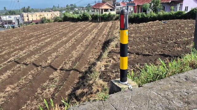 A wide shot showing a yellow and black road delineator pole slowly panning across a freshly tilled agricultural field in a rural hillside village.