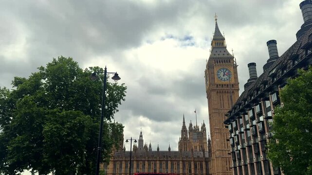 A red double decker bus passes a Gothic clock tower and government building under cloudy skies