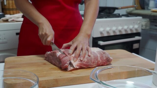 A person is cutting raw meat on a wooden cutting board in a kitchen setting, with a stove and countertop visible in the background. The scene is captured from a close-up, slightly elevated.