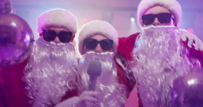Santa singers karaoke with microphone at disco party. Two friends link arms as another holds a disco ball, wearing sunglasses and beards under stage lights. Joyful party theme vibe idea.
