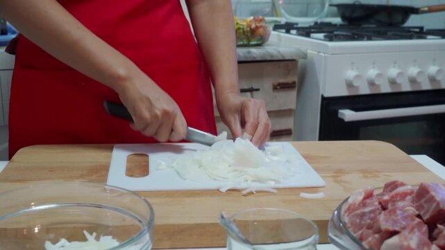 A person in a red apron is carefully chopping an onion on a wooden cutting board in a clean kitchen setting, viewed from a focused first-person perspective. The scene is vivid with rich red.