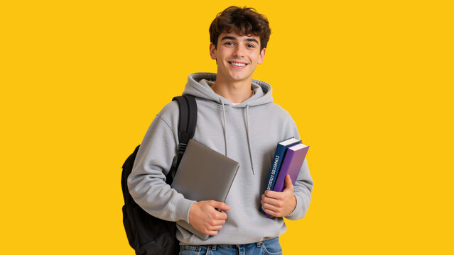 Smiling young male student holding laptop and books. Happy college guy with backpack in gray hoodie on yellow background. Copy space