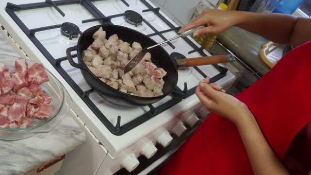 Cooking frying meat cubes, in kitchen stove, with frying pan, gas stove, meat cubes, glass bowl, from a over-the-shoulder view perspective, with red, white accents, and focused mood.