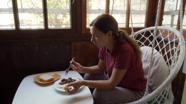 A calm woman eats lunch while sitting in a hanging chair at a small table. She enjoys a quiet, peaceful meal by the window. The wooden room feels cozy and serene.