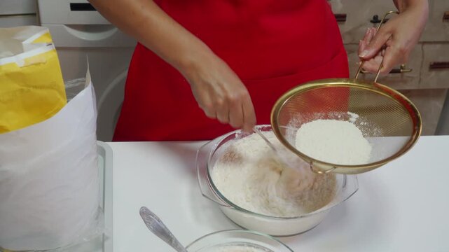 A passionate baker in a red apron shifts fresh flour into a mixing bowl on a white table. They create delicious dough with joyful, calm care. The bright kitchen feels warm and cozy.