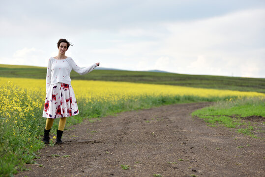 Woman hitchhiking on a dirt road next to a yellow canola flower field