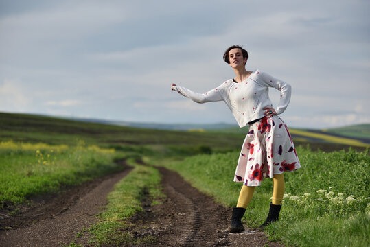 Woman hitchhiking on a dirt road next to a yellow canola flower field