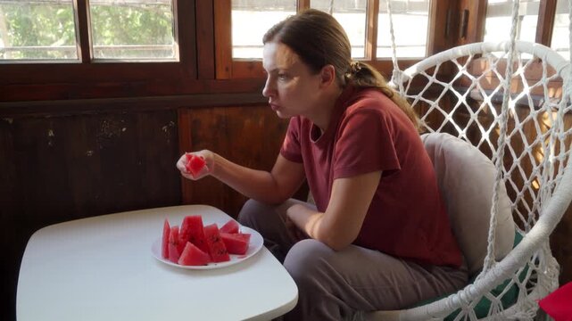 A relaxed woman eats fresh, sweet watermelon slices at a small table. She enjoys a calm, peaceful moment in her hanging chair. The sunlit wooden room feels cozy.