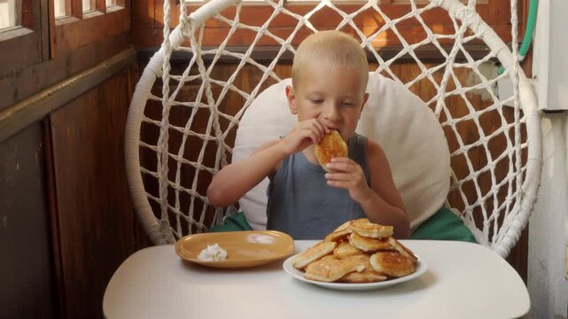 A happy boy joyfully eats fresh homemade pancakes while sitting in a cozy hanging chair on a sunny balcony. He looks excited and satisfied with his delicious breakfast.