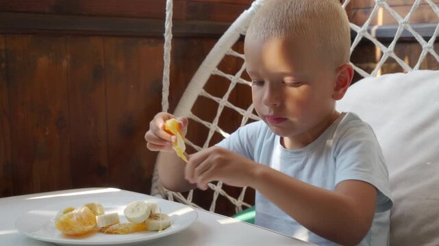 Close view of young boy concentrating as he peels tangerine and eats snack at table in cozy indoor room with soft natural light, capturing calm childhood lifestyle moment, with boy, child, tangerine