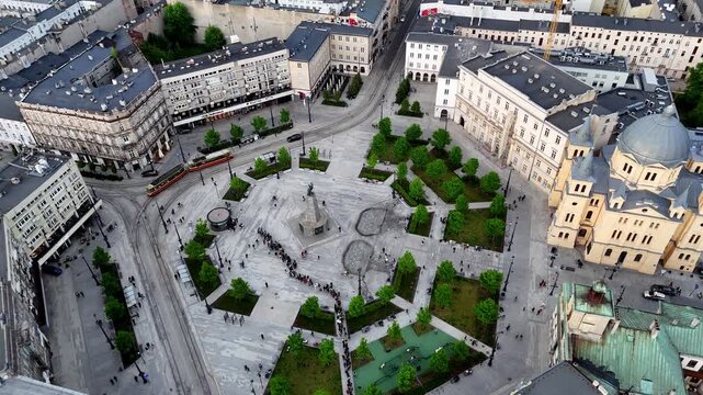 Drone video showing a high-angle aerial view of Plac Wolnosci in Lodz, Poland, featuring the Kosciuszko monument, Holy Spirit church, green patches, tram tracks, and historic buildings in daylight.
