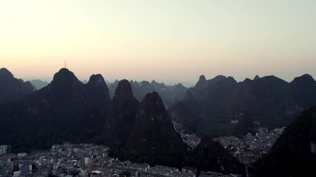 A panoramic view of distinctive karst mountain peaks framing a small town under a pale dusk sky, captured from a high vantage point.