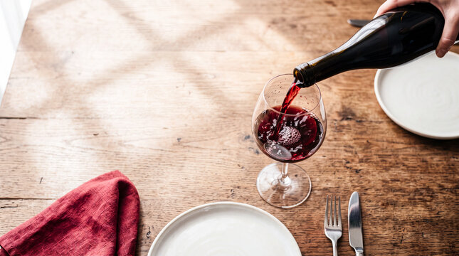 Red Wine Pour: A hand pours rich red wine into a crystal glass on a rustic wooden table, casting dappled sunlight and creating an intimate dining atmosphere.